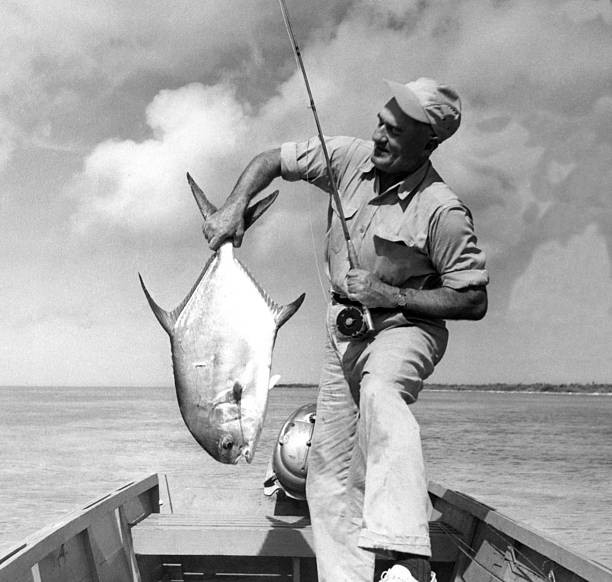 Joe Brooks stands a small wooden boat holding a permit caught fly - Old Photo