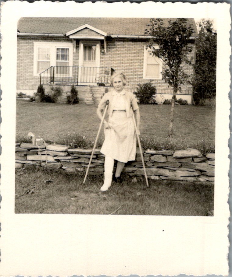 1950s Elderly Woman with Leg Cast Crutches at Brick Home Stone Wall Photograph