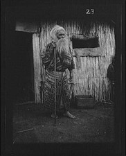 Photo:Ainu Man with Staff Outside Hut 1908 Village Thatched Structure picture